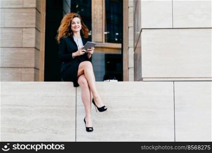 Beautiful elegant businesswoman in formal clothes and high-heeled shoes, having wavy hair and slender legs, using modern tablet for her work, looking at camera with happy look, smiling broadly