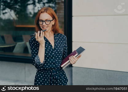 Beautiful elegant smiling business woman in dress using speakerphone or virtual voice assistant on mobile phone while standing with laptop in hand outdoors, records audio message for colleague. Beautiful smiling business woman using speakerphone or virtual voice assistant on mobile phone