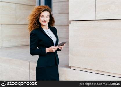 Beautiful elegant young businesswoman in black costume, having bushy curly hair and appealing appearance, holding tablet, reading news on Internet while posing against big builduing on street