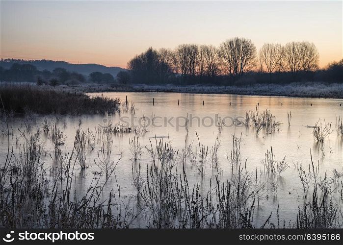 Beautiful English countryside lake image with frost and frozen lake in Winter at sunrise