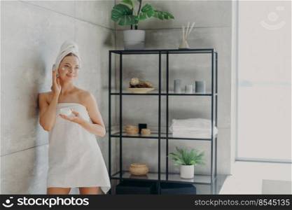 Beautiful European woman applies face mask, has flawless skin and looks somewhere thoughtfully, uses beauty cosmetics, stands wrapped in towel at cozy bathroom. Young lady applies body cream