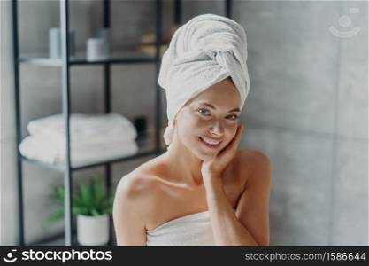 Beautiful European woman with makeup touches skin, has minimal makeup, has healthy glowing skin, wrapped in bath towel, enjoys rest at home. Spa woman poses in bathroom. Beauty, wellness concept