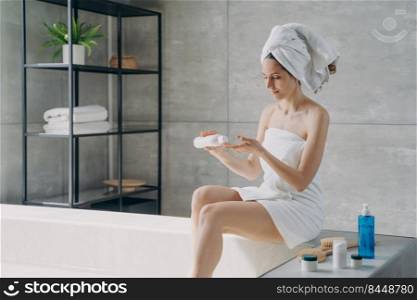 Beautiful european woman wrapped in towel applying body moisturizing cream after bathing. Young woman takes shower in morning at home. Anti-cellulite cosmetic, massage and bodycare.. Beautiful european woman wrapped in towel applying body moisturizing cream after bathing.