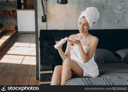 Beautiful european woman wrapped in towel is sitting in bedroom. Happy girl applying body moisturizing lotion squeezing it out from bottle after bathing. Morning shower at home.. Happy girl applying body moisturizing lotion squeezing it out from bottle after bathing at home.