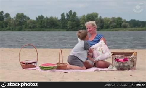 Beautiful excited mother and son having fun on picnic on the river bank during weekend and playing in pillow battle.