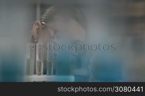Beautiful expirienced blonde female scientist in eyeglasses looking at test tubes with solution in chemical laboratory. Young attractive woman chemist working with test tubes in chemical research lab while conducting an experiment. Dolly shot.