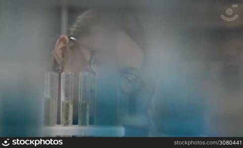 Beautiful expirienced blonde female scientist in eyeglasses looking at test tubes with solution in chemical laboratory. Young attractive woman chemist working with test tubes in chemical research lab while conducting an experiment. Dolly shot.