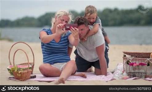 Beautiful family with child making a self portrait with a mobile phone during picnic at the beach. Woman with smartphone sitting on blanket and making selfie of her family. Father giving his excited son piggyback.