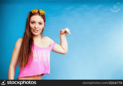 Beautiful fashion girl long hair in summer clothes sunglasses pointing copy space for product or text. Studio shot on blue background