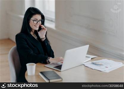 Beautiful female executive makes remote job, talks via mobile phone and checks information in laptop computer poses at workplace surrounded by coffee paper documents notepad has telephone conversation