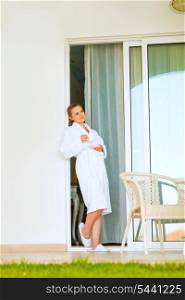 Beautiful female in bathrobe standing on terrace near big windows and having cup of coffee&#xA;