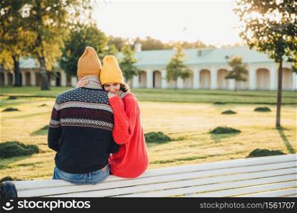 Beautiful female in loose red sweater embraces her boyfriend, missed him too much as didn`t see long ago, sit on bench in park, have pleasant conversation, admire wonderful landscape or nature