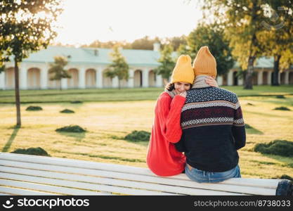 Beautiful female in loose red sweater embraces her boyfriend, missed him too much as didn`t see long ago, sit on bench in park, have pleasant conversation, admire wonderful landscape or nature