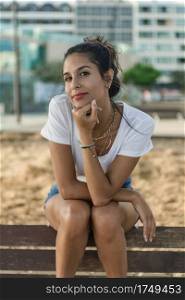 Beautiful female in trendy outfit holding head on fist and looking at camera while sitting on bench on blurred background of street. Stylish woman sitting on street
