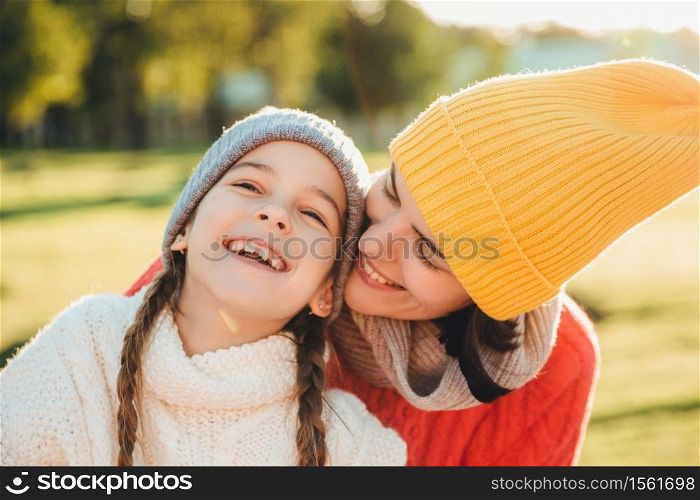 Beautiful female in yellow knitted hat kisses her adorable little child, have wonderful unforgettable time together. Small child smiles happily while recieves kiss from mother. Family relationships