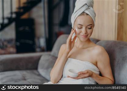 Beautiful female model enjoys softness of skin afrer taking bath, applies moisturizing cream on cheek, wears minimal makeup, wrapped in towel, sits on sofa alone, undergoes beauty treatments