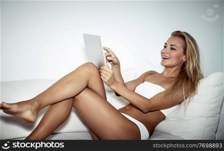 Beautiful female sitting on sofa with tablet at home, watching something or communicating with someone and laughing, enjoying day off, modern life of young people