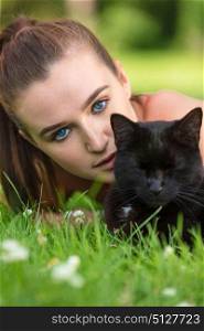 Beautiful female teenager girl young woman with blue eyes, laying down outside on grass with a black cat