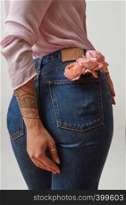 Beautiful female wearing in jeans with fresh roses of living coral color in a back pocket and pink shirt on a light gray background. Place for text. Concept of Woman's or Mother's Day.. Young pretty woman dressed in blue jeans and pink jacket with fresh roses in a back pocket and her hand tattoo on a gray background, copy space.