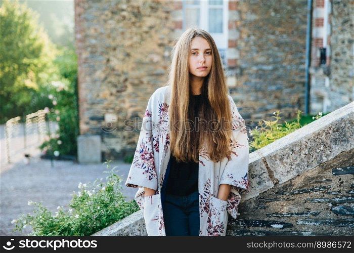 Beautiful female with long straight hair wears fashionable cape, stands over ancient city background, has excursion, goes sightseeing, poses near old builduings outdoor. People, lifestyle concept.