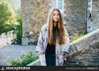 Beautiful female with long straight hair wears fashionable cape, stands over ancient city background, has excursion, goes sightseeing, poses near old builduings outdoor. People, lifestyle concept.