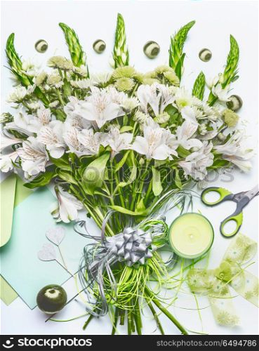 Beautiful festive bouquet with green flowers on white desk background with envelope and paper card mock up, ribbon, candle and scissors, top view. Holidays greeting concept, flat lay