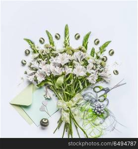Beautiful festive bouquet with green flowers on white desk background with envelope and paper card mock up, ribbon and scissors, top view. Holidays greeting concept