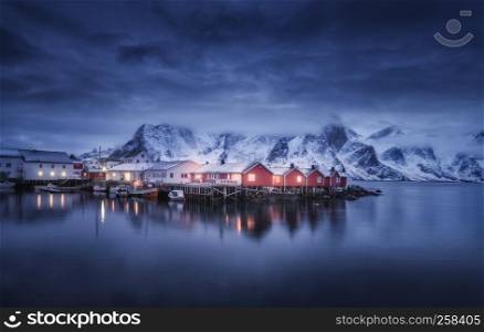Beautiful fishing village with boats at night, Lofoten islands, Norway. Winter landscape with houses, illumination, snowy mountains, sea, blue cloudy sky reflected in water at dusk. Norwegian rorbuer