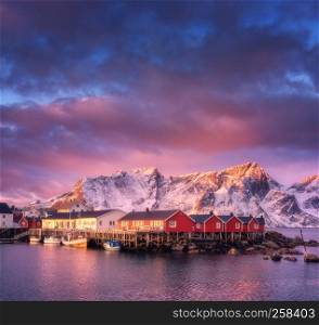 Beautiful fishing village with boats at sunrise, Lofoten islands, Norway. Winter landscape with houses, snowy mountains, sea, boats, colorful sky with clouds. Norwegian traditional red rorbu and rocks