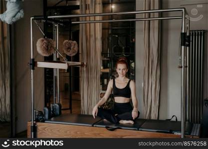 Beautiful fit woman sitting in very relaxed lotus position on top of trapeze table or Cadillac reformer, smiling and feeling satisfied after stretching workout in studio. Concept of pilates and sport. Young athletic woman sitting in very relaxed lotus position on top of pilates machine