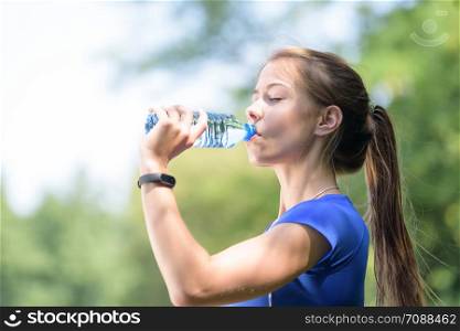 Beautiful fitness girl drinks mineral water from a bottle in a park on a sunny day in close-up