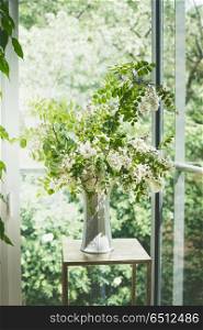 Beautiful flowers bunch with blossom acacia branches in white vase in living room at window. Interior design and ideas . Still life