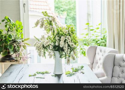Beautiful flowers bunch with blossom acacia branches in white vase on table in living room at window. Interior design and ideas . Still life