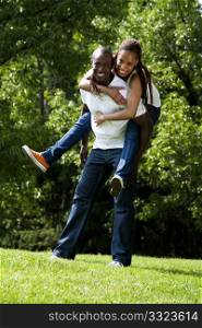 Beautiful fun happy smiling laughing African American couple piggyback playing in the park, woman hugging man, wearing white shirts and blue jeans.