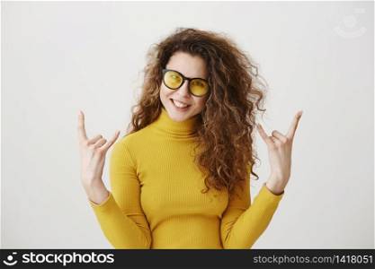 Beautiful funny amazed rocker girl with curly hairstyle and standing with rock sign and looking at camera. studio shot, isolated on grey background.. Beautiful funny amazed rocker girl with curly hairstyle and standing with rock sign and looking at camera. studio shot, isolated on grey background