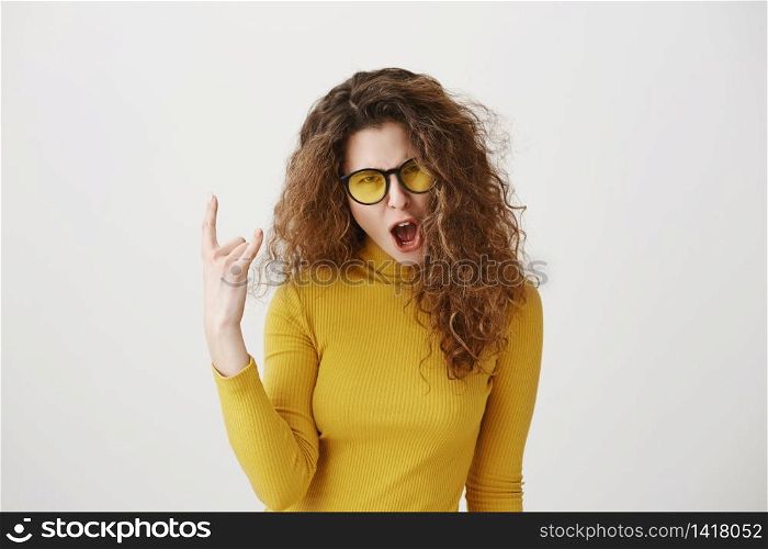 Beautiful funny amazed rocker girl with curly hairstyle and standing with rock sign and looking at camera. studio shot, isolated on grey background.. Beautiful funny amazed rocker girl with curly hairstyle and standing with rock sign and looking at camera. studio shot, isolated on grey background