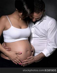 Beautiful gentle pregnant woman and her loving husband with tenderness touching and looking at belly over black background, young family and love concept