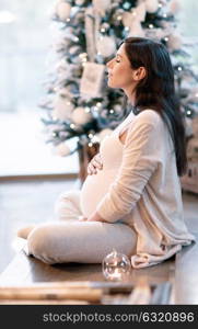 Beautiful gentle pregnant woman with closed eyes sitting near decorated Christmas tree and with love touching her belly, with pleasure spending winter holidays at home