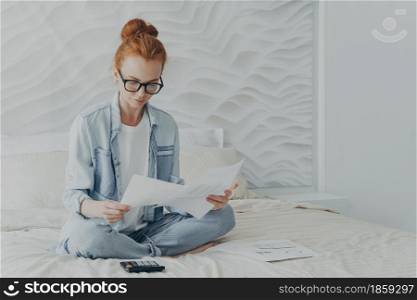 Beautiful ginger woman freelancer looks attentively at paper documents calculates on calculator manages finances at home does savings for future wears denim shirt and jeans sits crossed legs on bed. Beautiful ginger woman freelancer looks attentively at documents calculates manages finances