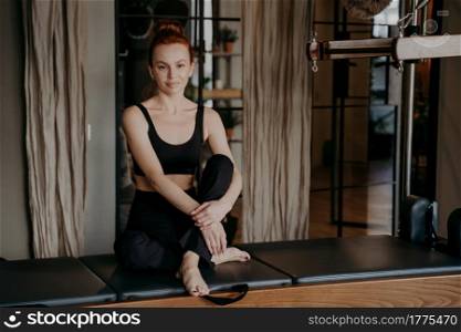 Beautiful ginger woman in sportswear sitting on Cadillac machine with arms around legs in calm and relaxed position after exercising in gym or fitness studio. Sport and healthy lifestyle. Beautiful young pilates trainer sitting with arms around legs in calm and relaxed position