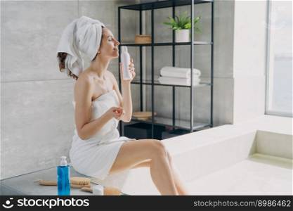 Beautiful girl enjoys smell of body lotion perfume and has rest in bathroom. Attractive caucasian woman wrapped in towel after bathing. Young lady takes shower at home. Tranquility and relaxation.. Girl enjoys smell of body lotion perfume and has rest in bathroom. Tranquility and relaxation.