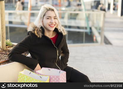 Beautiful girl holding shopping bags, looking at camera and smiling while sitting on a bench