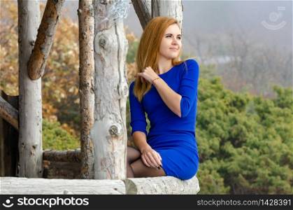 Beautiful girl in bright blue dress sits on a wooden terrace and looks into the distance