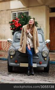 Beautiful girl in city. happy young woman standing near a retro car decorated with Christmas tree. Cold happy winter day. Holidays, christmas, winter, love, beauty concept.. Beautiful girl in city. happy young woman standing near a retro car decorated with Christmas tree. Cold happy winter day. Holidays, christmas, winter, love, beauty concept