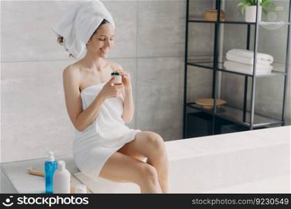 Beautiful girl is holding body cream jar and smiling. Gorgeous european woman wrapped in towel after bathing. Young woman takes shower at home. Morning body care, skincare in bathroom.. Beautiful girl is holding body cream jar and smiling. . Young woman takes shower at home.