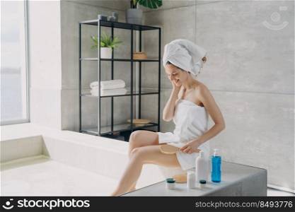 Beautiful girl is sitting on bathtub and making body massage with brush. European lady wrapped in towel after bathing. Young smiling woman takes shower in bathroom at home. Morning freshness.. Beautiful girl is sitting on bathtub and making body massage with brush. Morning freshness.
