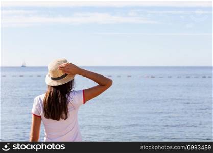 Beautiful girl looks at the sea. Young girl in a hat looking at a calm sea and blue skies back view.. Beautiful girl looks at the sea. Young girl in a hat