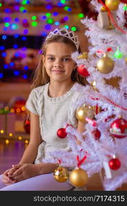 Beautiful girl sits at the Christmas tree against the background of blurred lights