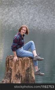 beautiful girl sits on a stump and smiling at the camera on a background of a mountain lake