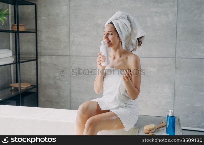 Beautiful girl takes a smell of body cream and relaxing in bathroom. Attractive caucasian woman wrapped in towel after bathing. Lovely young lady takes shower. Cosmetics applying and skin care.. Beautiful girl takes smell of body cream and relaxing in bathroom. Cosmetics applying and skin care.